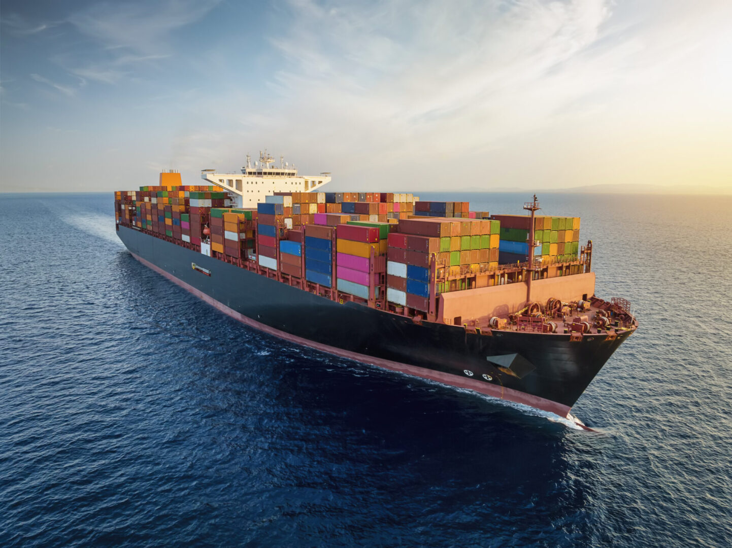 Aerial view of a large, loaded container cargo ship traveling over open ocean during sunset time